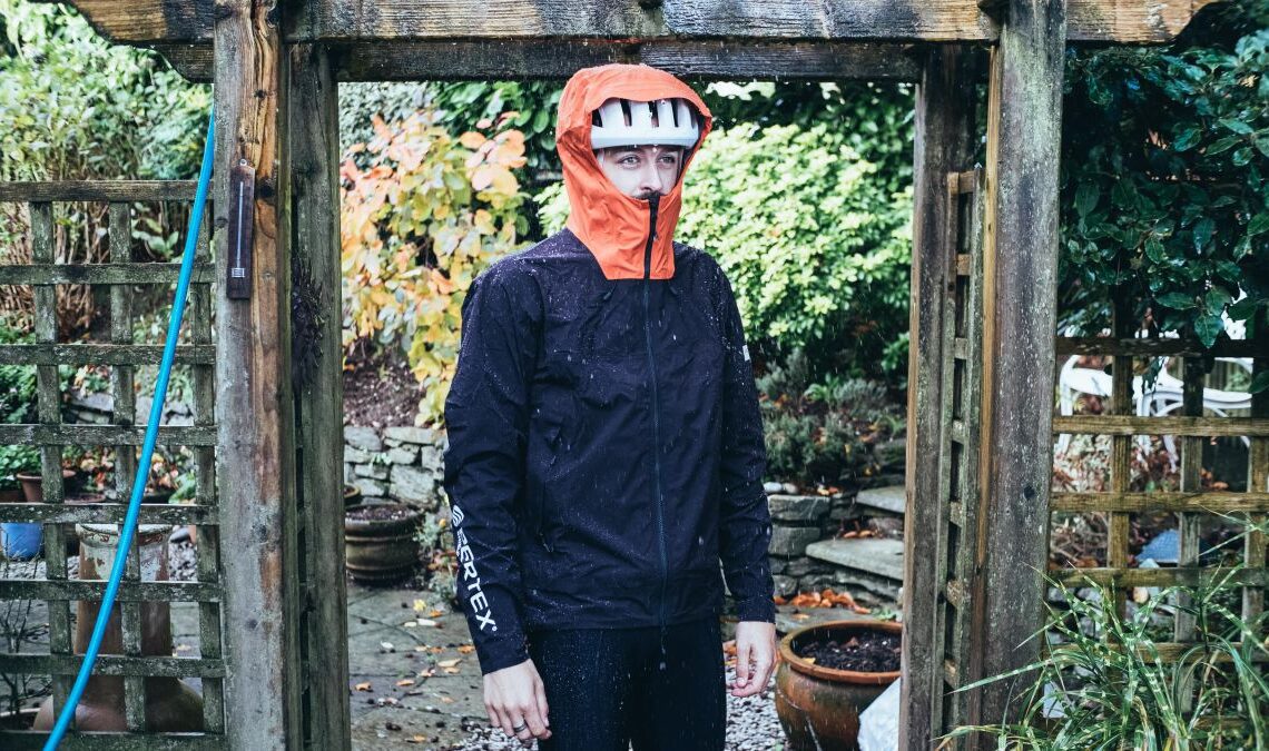 A white man wears a waterproof jacket under a stream of water in a garden