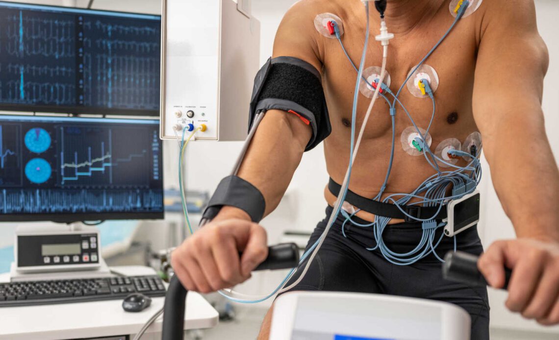 Photo of an unrecognizable male athlete on an exercise bike and LCD monitors showing biometric data during biometric testing in a lab.