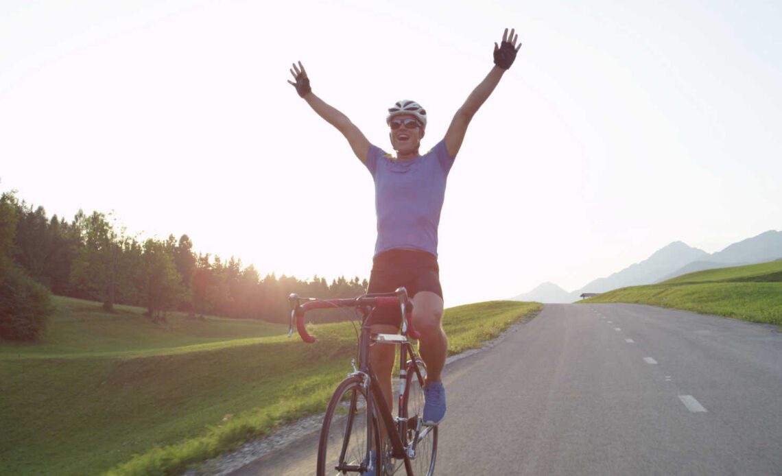 Professional cyclist raising his arms victoriously after winning race. Cheerful biking athlete celebrates victory in quiet countryside. Pro cyclist cheerfully rides road bike with no hands.